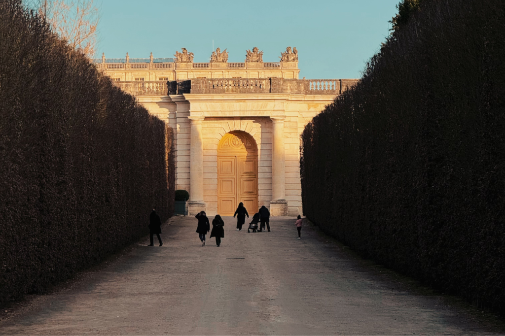 Visitors walking towards a sunlit entrance at the Palace of Versailles, surrounded by manicured hedges, during golden hour.