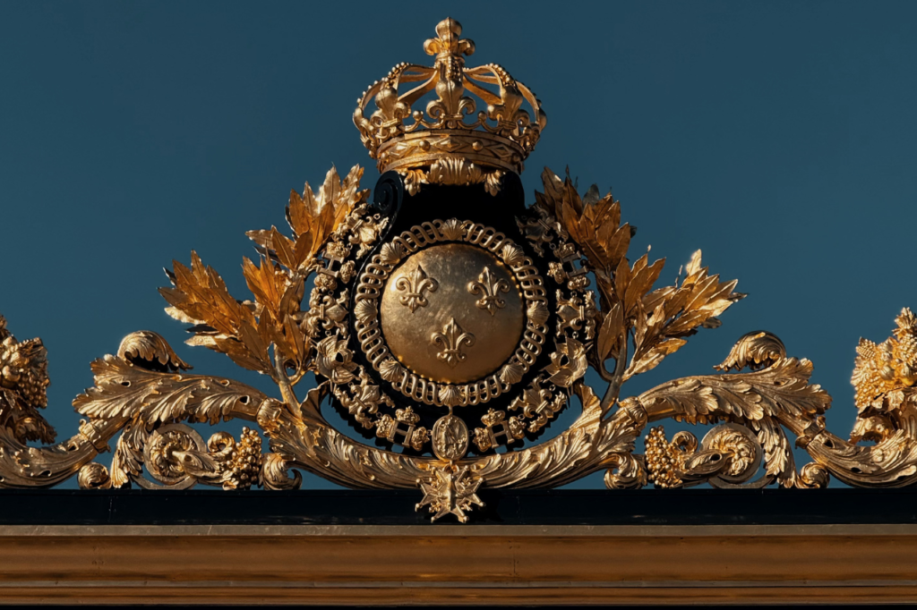 A close-up of the golden royal crest at the Palace of Versailles, shining under the sunlight against a deep blue sky.